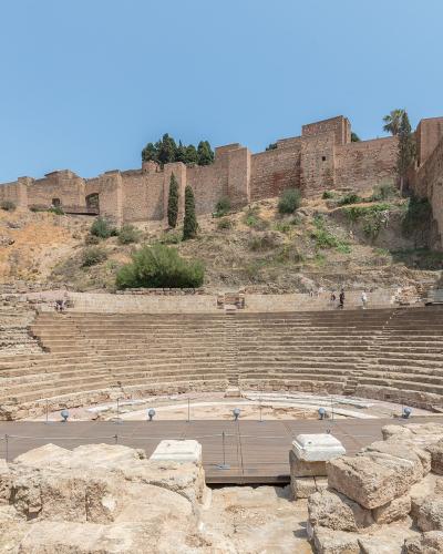 The image shows the stone seating tiers of an ancient Roman theatre with a modern wooden stage platform in the foreground, set against a hillside topped by the Alcazaba fortress walls.