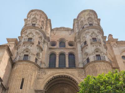 A low-angle photograph of the ornate stone facade of a large cathedral featuring two prominent, rounded corner towers with balconies and a central arched entrance, set against a clear blue sky with green foliage in the foreground.