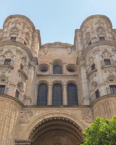 A low-angle photograph of the ornate stone facade of a large cathedral featuring two prominent, rounded corner towers with balconies and a central arched entrance, set against a clear blue sky with green foliage in the foreground.