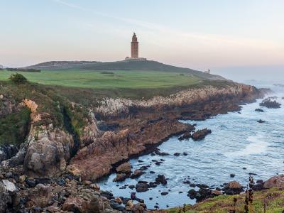 A coastal landscape featuring a prominent ancient Roman lighthouse situated on a grassy promontory overlooking the ocean, with rocky cliffs in the foreground and a city skyline visible in the distance to the left.
