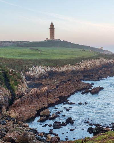 A coastal landscape featuring a prominent ancient Roman lighthouse situated on a grassy promontory overlooking the ocean, with rocky cliffs in the foreground and a city skyline visible in the distance to the left.