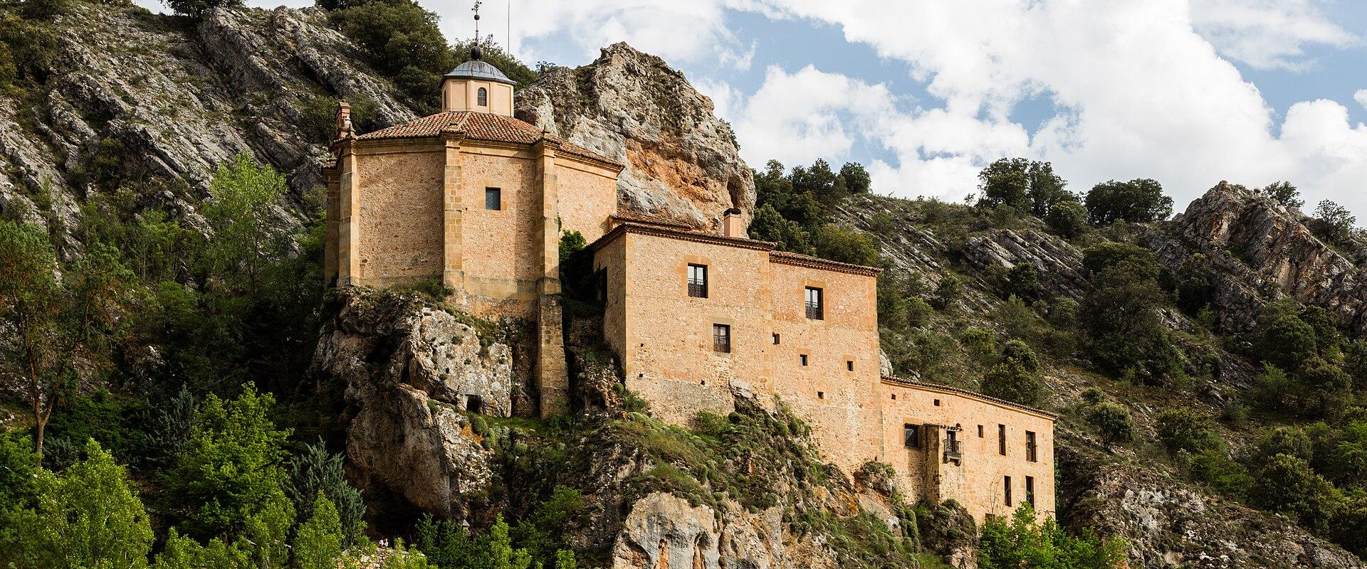 A stone religious building complex built directly into a rocky cliff face next to a river, featuring a small octagonal chapel with a dome and a cross on top.
