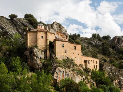 A stone religious building complex built directly into a rocky cliff face next to a river, featuring a small octagonal chapel with a dome and a cross on top.