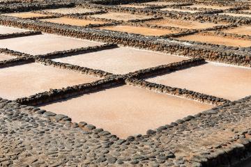 A close-up view of rectangular evaporation ponds lined with dark volcanic stones, containing pinkish saltwater and crystallizing salt.