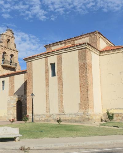 A beige and brick church with a bell tower, set in a town square with benches and lampposts under a blue sky.