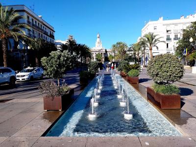 A long, narrow reflecting pool with multiple water jets runs through a paved pedestrian plaza lined with palm trees and large planters, with white buildings and a prominent church tower visible in the background.