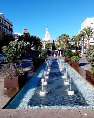 A long, narrow reflecting pool with multiple water jets runs through a paved pedestrian plaza lined with palm trees and large planters, with white buildings and a prominent church tower visible in the background.