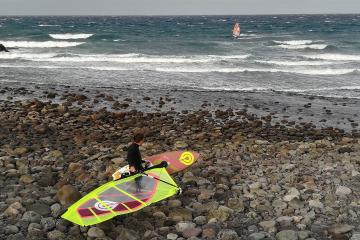 A windsurfer sits on a rocky beach preparing their gear, with another windsurfer visible in the ocean waves in the background.