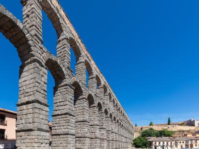 A low-angle view of a massive, two-tiered Roman stone aqueduct with numerous arches, set against a clear blue sky with people walking in the plaza below.