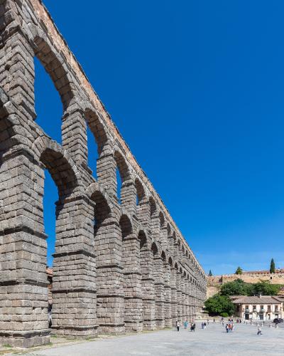 A low-angle view of a massive, two-tiered Roman stone aqueduct with numerous arches, set against a clear blue sky with people walking in the plaza below.