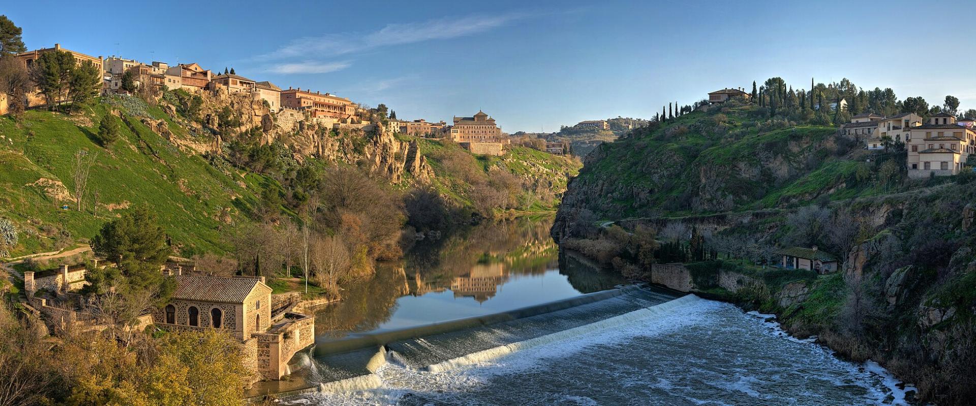 A panoramic view of a historic city built on steep cliffs overlooking a river with a weir and a small stone building in the foreground.