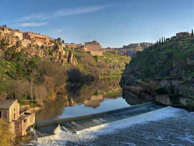 A panoramic view of a historic city built on steep cliffs overlooking a river with a weir and a small stone building in the foreground.