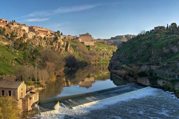 A panoramic view of a historic city built on steep cliffs overlooking a river with a weir and a small stone building in the foreground.