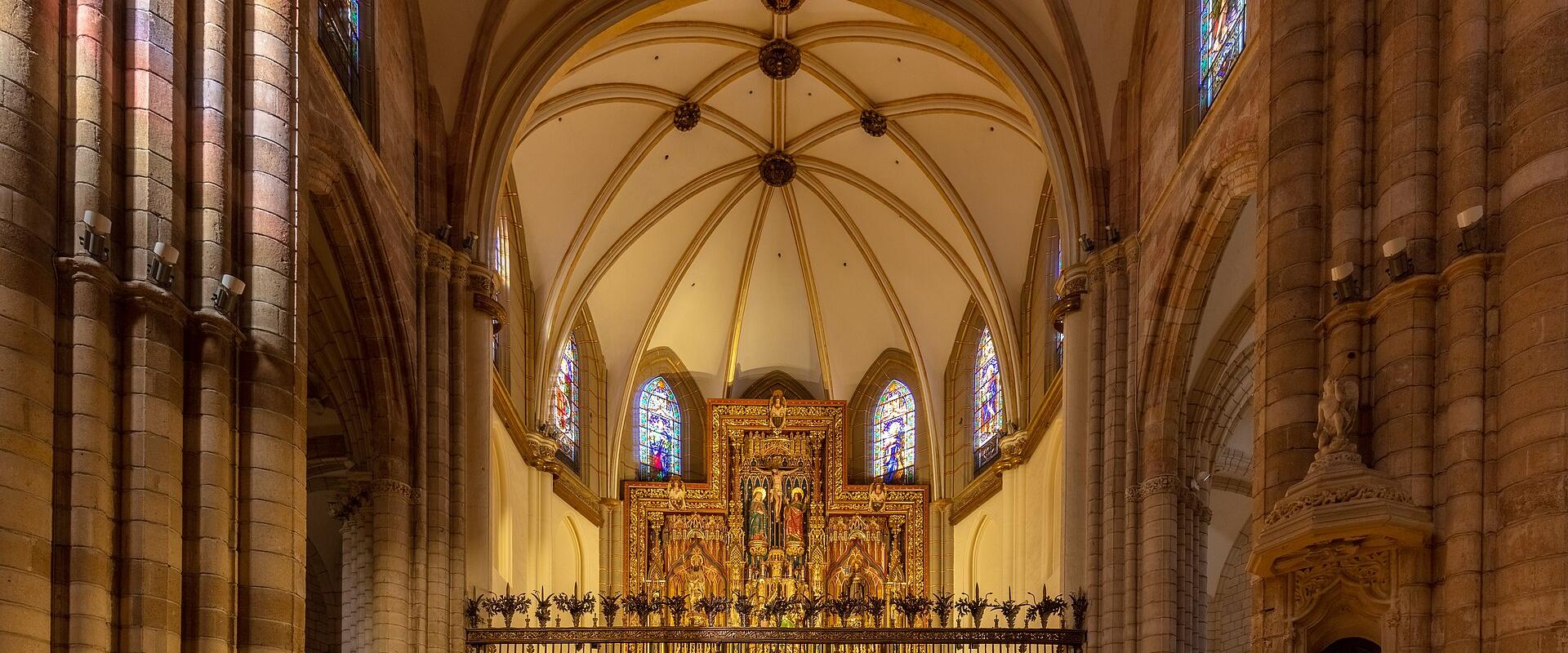 The interior of a large Gothic cathedral featuring ribbed vaults, tall stone columns, stained glass windows, and an ornate golden altarpiece behind a metal screen.