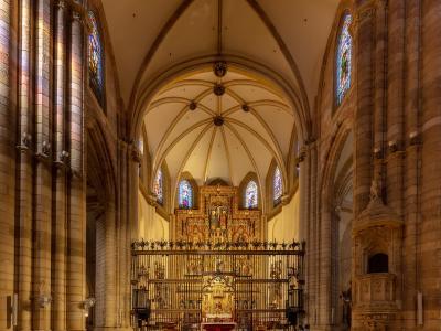 The interior of a large Gothic cathedral featuring ribbed vaults, tall stone columns, stained glass windows, and an ornate golden altarpiece behind a metal screen.