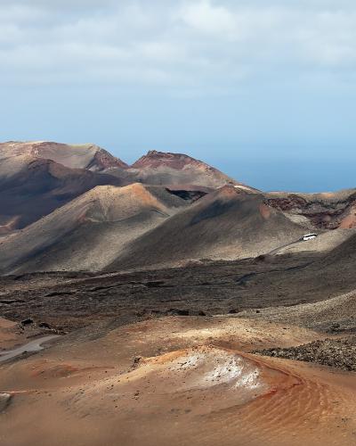 A panoramic view of the Timanfaya National Park landscape in Lanzarote, featuring multi-colored volcanic cones, black lava fields, and a winding road with a small white vehicle.