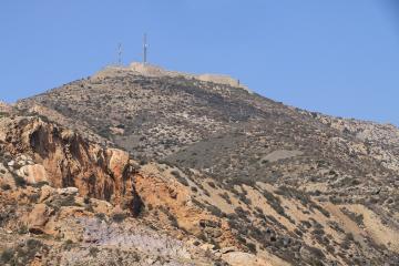 A stone fortress and communication towers atop a dry, rocky hill under a clear blue sky.