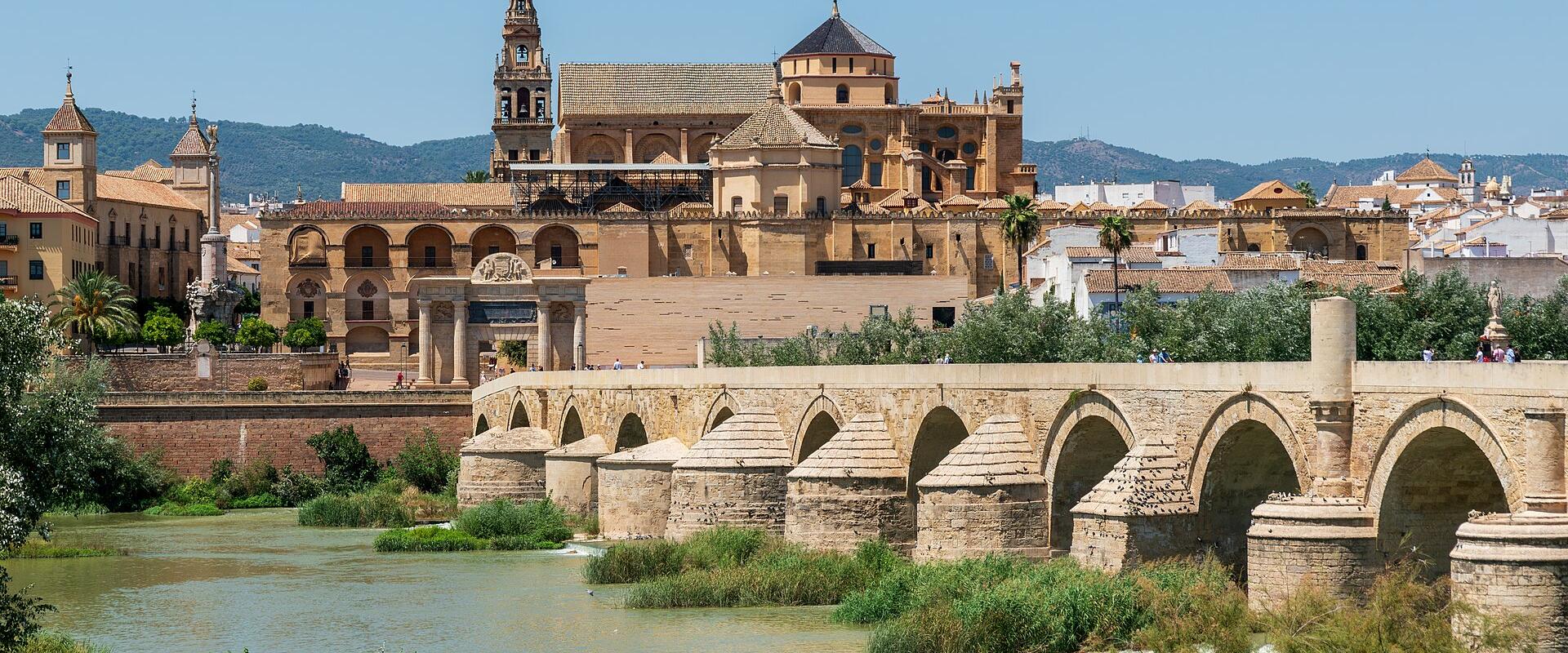 A stone Roman bridge with multiple arches spans a river in the foreground, with the Mezquita-Catedral (a large mosque-cathedral complex with a bell tower and dome) and other historic buildings visible on the riverbank under a clear blue sky.