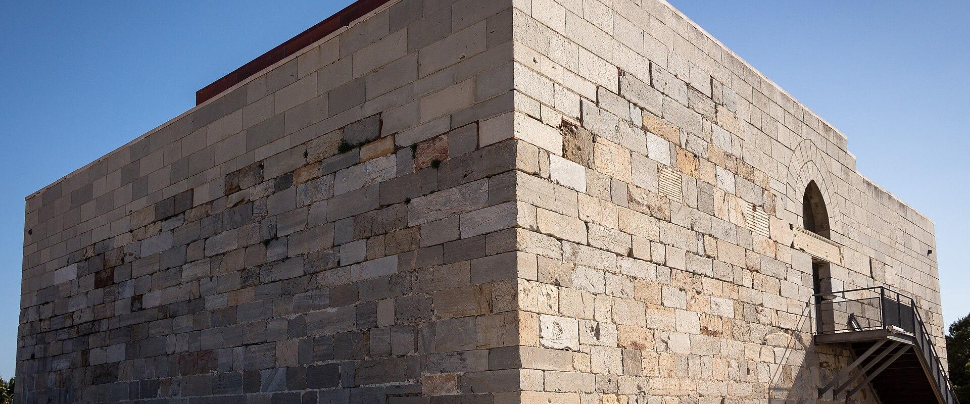 A low-angle shot of a large, rectangular stone fortress building with crenellated battlements, set against a clear blue sky on a gravel surface.