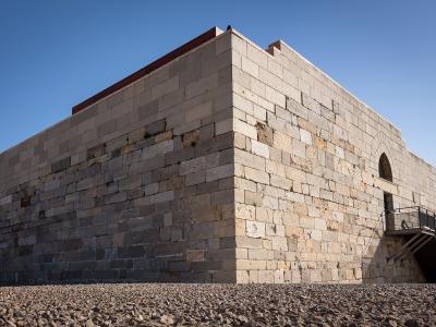 A low-angle shot of a large, rectangular stone fortress building with crenellated battlements, set against a clear blue sky on a gravel surface.
