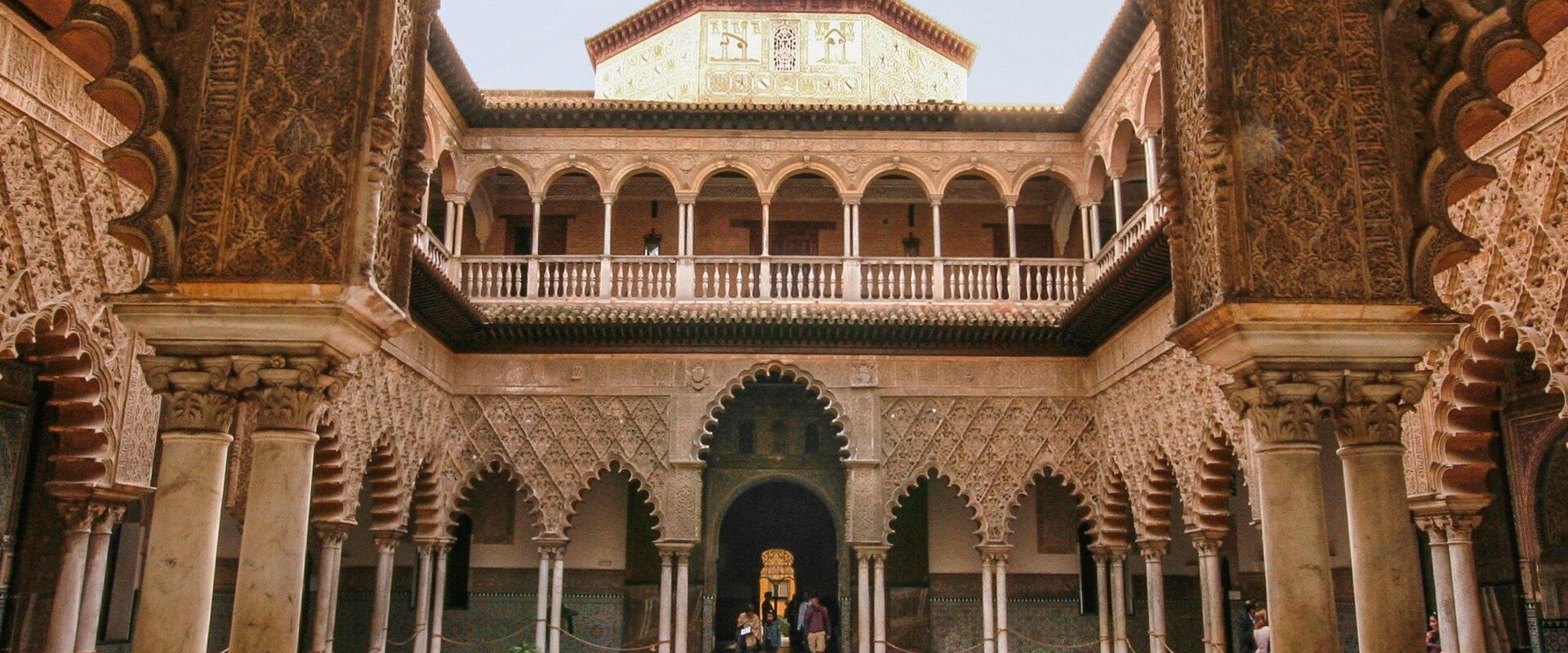 A close-up photograph of an intricately carved stone capital on a pillar, featuring Moorish-style arabesque patterns and Arabic calligraphy, set within an arched corridor.