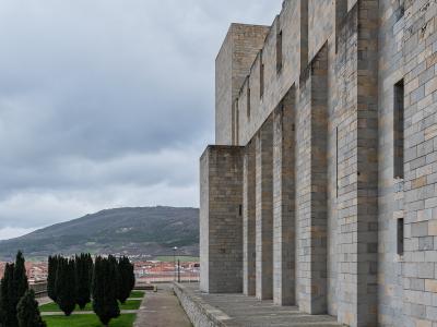 A modern stone building with a colonnade of large rectangular pillars, adjacent to a grassy area with cypress trees and a view of a town and hill in the distance.