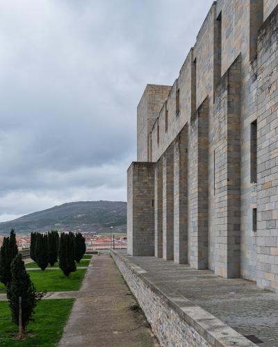 A modern stone building with a colonnade of large rectangular pillars, adjacent to a grassy area with cypress trees and a view of a town and hill in the distance.