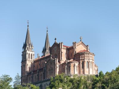A large, ornate basilica with two prominent spires and a red-tiled roof, situated atop a steep, forested hill under a clear blue sky.