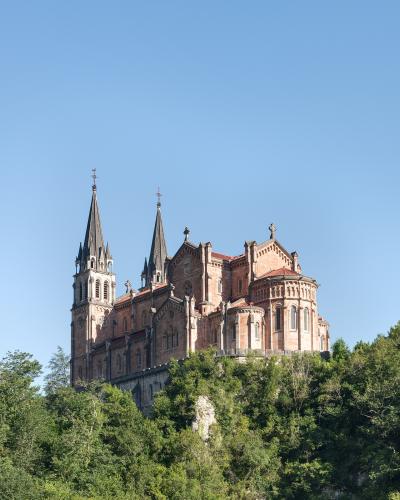 A large, ornate basilica with two prominent spires and a red-tiled roof, situated atop a steep, forested hill under a clear blue sky.