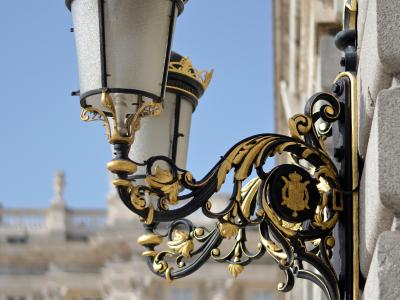 An ornate black and gold wall-mounted lantern with a crown finial and a royal coat of arms emblem, attached to a stone building facade with a blurred classical palace in the background.