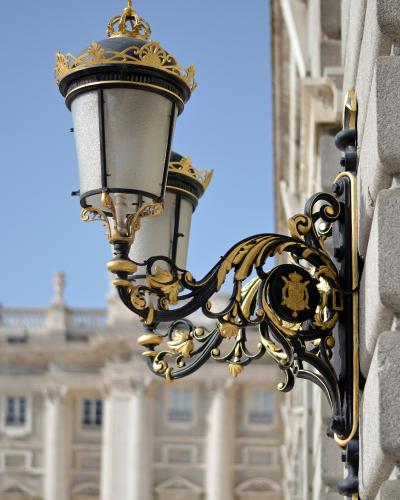 An ornate black and gold wall-mounted lantern with a crown finial and a royal coat of arms emblem, attached to a stone building facade with a blurred classical palace in the background.