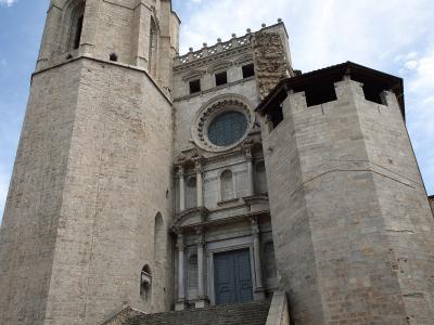 A low-angle photograph of a large stone church featuring a tall, square bell tower with Gothic-style belfry openings, a Baroque-style main facade with a rose window, and a wide stone staircase leading up to the entrance, flanked by a defensive-lookin