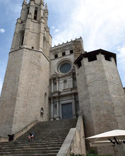 A low-angle photograph of a large stone church featuring a tall, square bell tower with Gothic-style belfry openings, a Baroque-style main facade with a rose window, and a wide stone staircase leading up to the entrance, flanked by a defensive-lookin