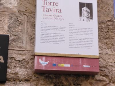A close-up photograph of an informational sign mounted on a rough stone wall, featuring the text "Torre Tavira" and "Cámara Oscura / Camera Obscura" along with a small illustration of the tower and logos for Cádiz.