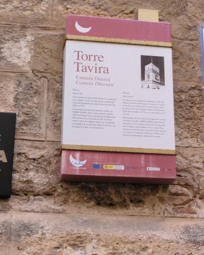 A close-up photograph of an informational sign mounted on a rough stone wall, featuring the text "Torre Tavira" and "Cámara Oscura / Camera Obscura" along with a small illustration of the tower and logos for Cádiz.