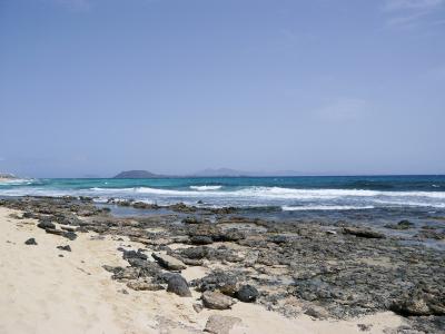 A rocky shoreline with sand in the foreground, turquoise ocean waves, and a distant island silhouette under a clear blue sky.