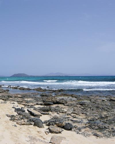 A rocky shoreline with sand in the foreground, turquoise ocean waves, and a distant island silhouette under a clear blue sky.