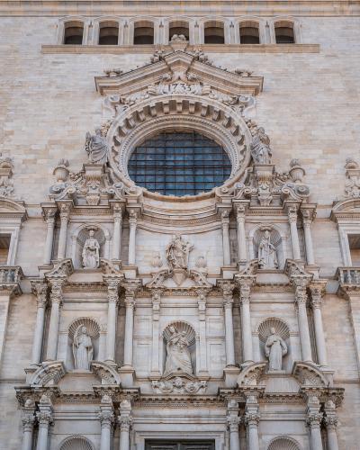 A close-up view of the ornate, light-colored stone Baroque facade of a large cathedral, featuring a central circular window, multiple statues in niches, and the date "1763" inscribed above the window.
