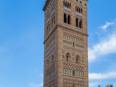 A tall, ornate brick bell tower with intricate geometric tilework and arched openings, standing next to a lower church building and a large wrought-iron cross in a town square.