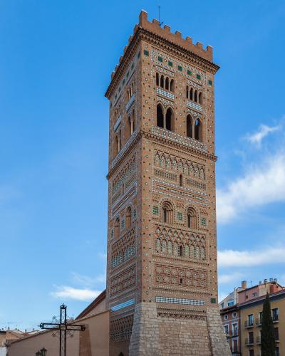 A tall, ornate brick bell tower with intricate geometric tilework and arched openings, standing next to a lower church building and a large wrought-iron cross in a town square.