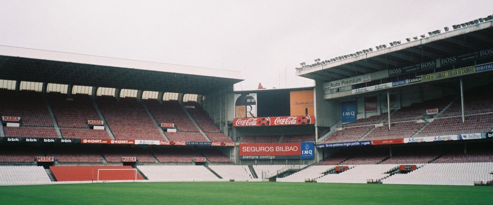 The image shows the interior of a football stadium with red and white seating, advertising hoardings for "Seguros Bilbao" and "Coca-Cola," and a large scoreboard structure.