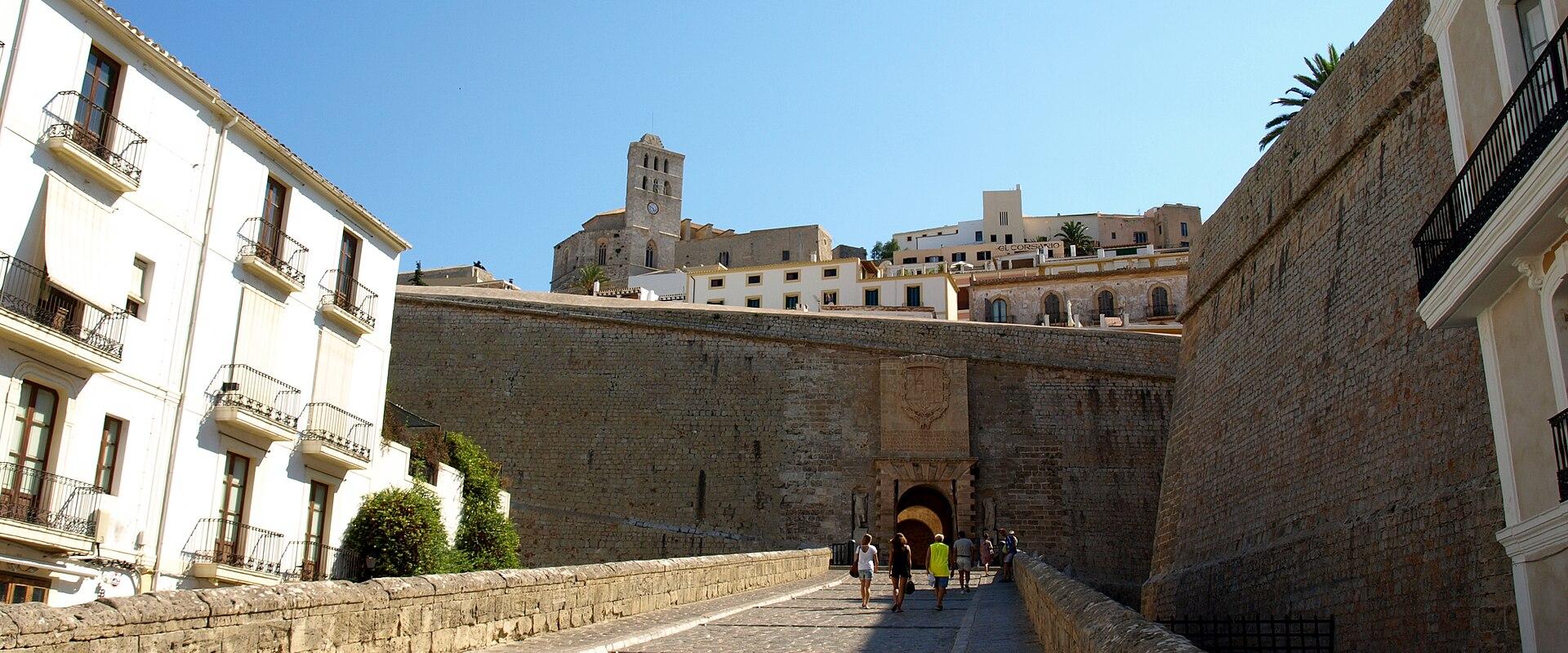 A stone ramp leading up to a fortified gateway with a coat of arms above the arch, flanked by white buildings and a high stone wall, with a church tower visible in the background.