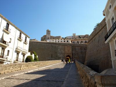 A stone ramp leading up to a fortified gateway with a coat of arms above the arch, flanked by white buildings and a high stone wall, with a church tower visible in the background.