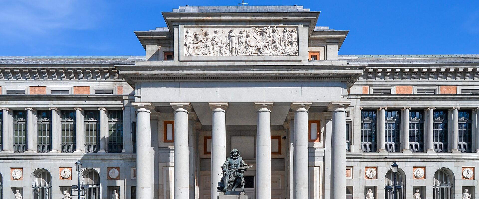 The image shows the neoclassical facade of a large museum building with a central portico supported by six columns, a Spanish flag flying above, and a bronze statue of Miguel de Cervantes standing on a pedestal in the foreground.