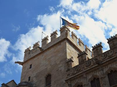 A low-angle view of a Gothic stone tower with crenellated battlements, a gargoyle, and a flag flying from the top against a blue sky.