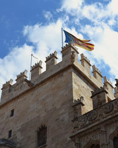 A low-angle view of a Gothic stone tower with crenellated battlements, a gargoyle, and a flag flying from the top against a blue sky.
