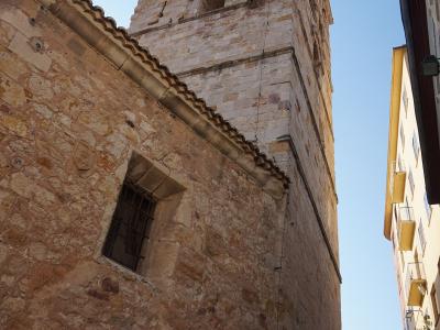 A low-angle view of a tall, rectangular stone bell tower with arched openings near the top, attached to a rougher stone building with a tiled roof and a barred window, set against a blue sky with adjacent modern buildings.