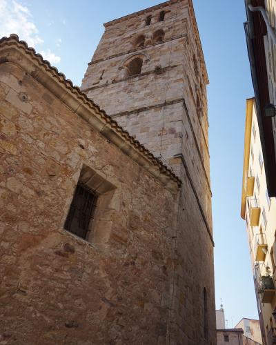 A low-angle view of a tall, rectangular stone bell tower with arched openings near the top, attached to a rougher stone building with a tiled roof and a barred window, set against a blue sky with adjacent modern buildings.