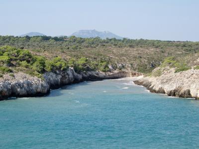 A coastal inlet with turquoise water, rocky cliffs, and a small sandy beach at the end, backed by green scrubland and distant hills.