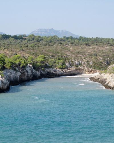 A coastal inlet with turquoise water, rocky cliffs, and a small sandy beach at the end, backed by green scrubland and distant hills.